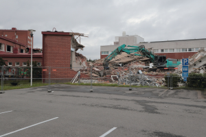 Großes Gebäude in einem Parkplatz mit einem Bagger, Metallstangen, Zaun, Schild, Pflanzen, Gras, Bäumen, benachbarten Gebäuden und einem bewölkten Himmel abgerissen.
