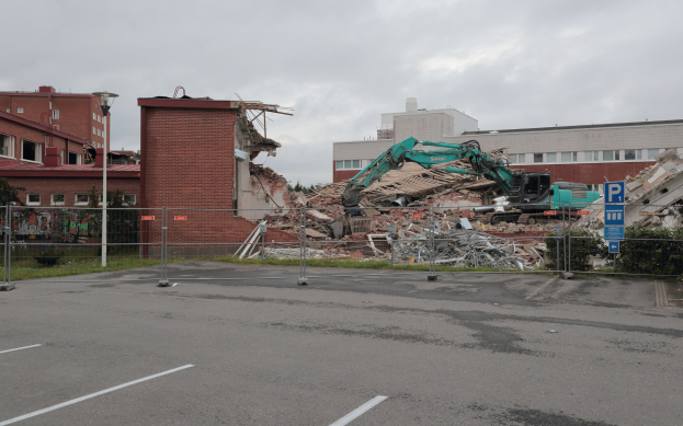 Großes Gebäude in einem Parkplatz mit einem Bagger, Metallstangen, Zaun, Schild, Pflanzen, Gras, Bäumen, benachbarten Gebäuden und einem bewölkten Himmel abgerissen.