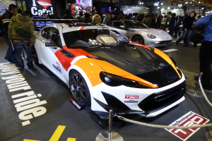 Eine Gruppe von Menschen betrachtet ein Rennfahrzeug auf der Tokyo Motor Show, umgeben von Absperrpollern mit Seilen und Text/Bildtafeln im Hintergrund unter beleuchteter Decke.