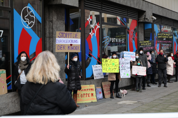 Eine Gruppe von Menschen mit Masken und Schildern protestiert vor einem Glasfrontgebäude, mit Texttafeln, Kameras und einem Hund.