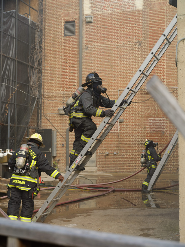 Feuerwehrleute in Helmen und Lufttanks, die eine Leiter vor einem Backsteingebäude hochklettern, mit Rohren auf dem Boden und einer Metallstange am unteren Ende, mit einem weiteren Gebäude mit Fenstern und einem Netz im Hintergrund.