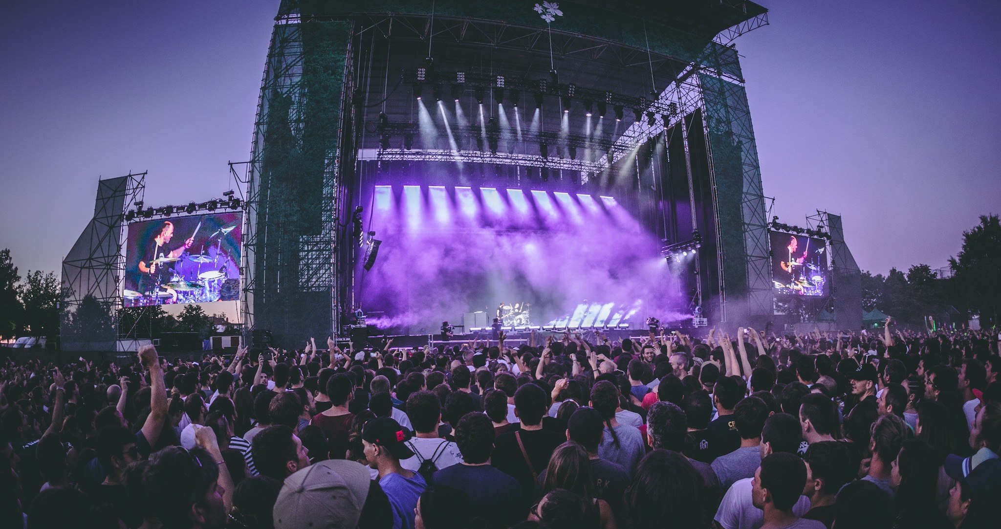 Eine große Menge bei einem Musikfestival schaut auf der Bühne mit Instrumenten und Bildschirmen, die vor einem Gebäude, Bäumen und einem blauen Himmel aufgebaut sind.