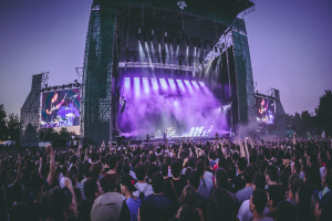 Eine große Menge bei einem Musikfestival schaut auf der Bühne mit Instrumenten und Bildschirmen, die vor einem Gebäude, Bäumen und einem blauen Himmel aufgebaut sind.