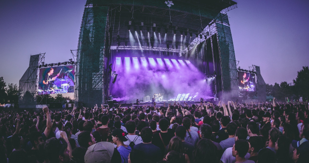 Eine große Menge bei einem Musikfestival schaut auf der Bühne mit Instrumenten und Bildschirmen, die vor einem Gebäude, Bäumen und einem blauen Himmel aufgebaut sind.