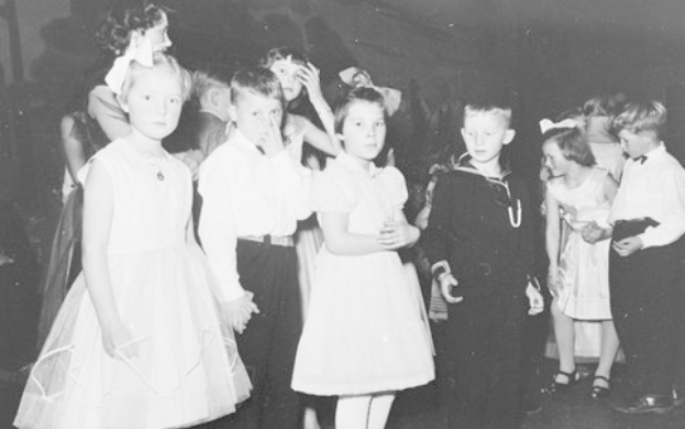 Schwarzes und weißes Bild einer Gruppe von Kindern, die auf einer Tanzfläche stehen, lächeln und glücklich aussehen, mit einer Wand im Hintergrund.