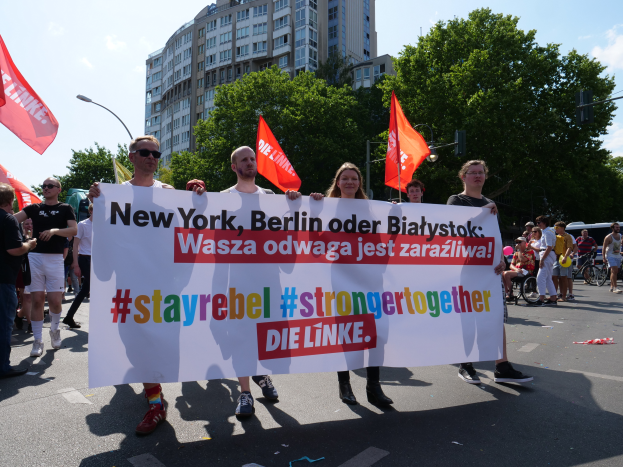 Eine Gruppe von Menschen geht eine von Bäumen gesäumte Straße in Berlin, Deutschland, entlang und hält ein Banner mit der Aufschrift "Stay Rebel Stronger Together" in der Hand, während Gebäude und Laternenpfähle sowie ein bewölkter Himmel zu sehen sind.