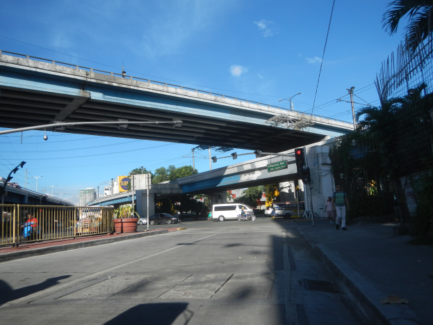 Eine Stadtstraße mit einer Fußgängerbrücke, Fahrzeugen, Fußgängern, einem Zaun, Topfpflanzen, Schildern, Laternen, Strommasten mit Drähten, Bäumen, Gebäuden und einem bewölkten Himmel.