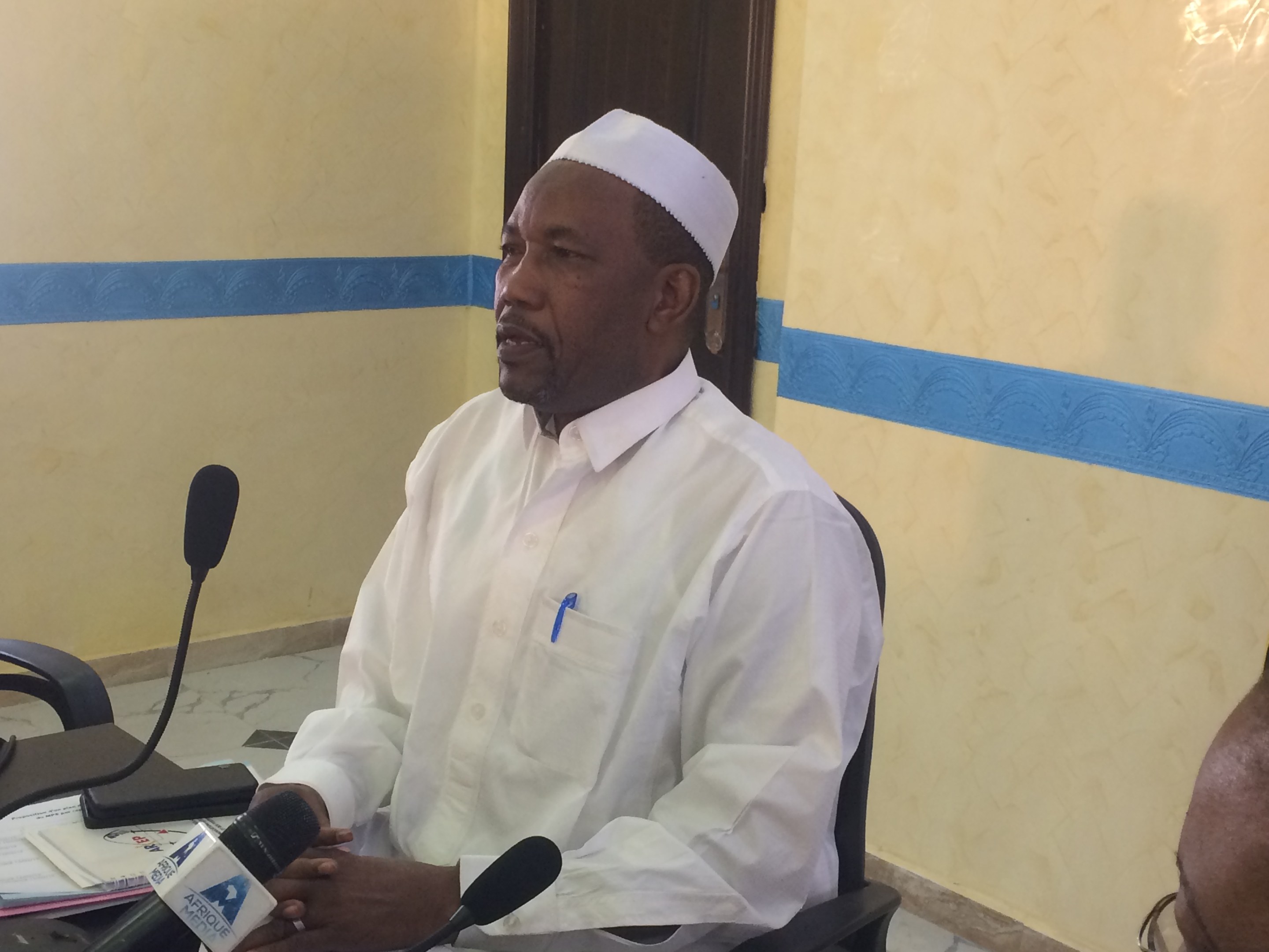 Ein Mann in weißem Anzug und Hut sitzt an einem Tisch mit Mikrofonen und spricht, während Papiere und Gegenstände auf dem Tisch liegen; rechts von ihm sitzt eine Person mit Brille, im Hintergrund eine Tür und eine Wand.