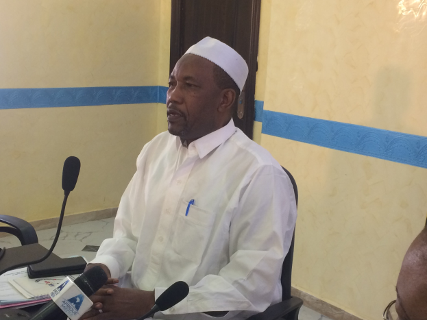 Ein Mann in weißem Anzug und Hut sitzt an einem Tisch mit Mikrofonen und spricht, während Papiere und Gegenstände auf dem Tisch liegen; rechts von ihm sitzt eine Person mit Brille, im Hintergrund eine Tür und eine Wand.