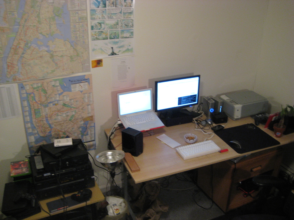 Ein Raum mit einem Tisch, auf dem ein Laptop, ein Lautsprecher, ein Monitor, eine Tastatur, ein Ball, eine Maus, ein Scanner und Papiere liegen, sowie ein weiterer Tisch mit Elektronikgeräten, mit Charts und Postern an der Wand dahinter.