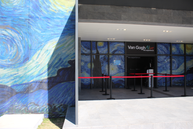 Der Eingang eines Gebäudes zeigt ein großes Wandgemälde, ein Namensschild mit der Aufschrift "Van Gogh Alive", Absperrpoller mit Bändern, Gras auf dem Boden und Lampen an der Decke.