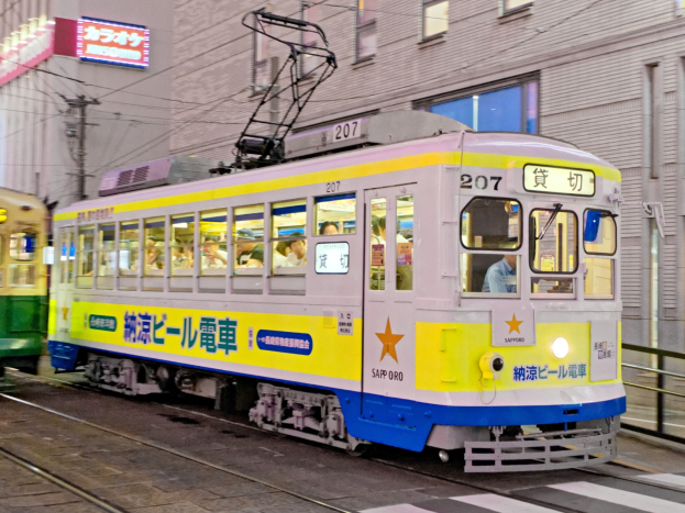 Eine gelbe und blaue Straßenbahn auf einer Stadtstraße bei Nacht mit Menschen darin, Gebäude mit Fenstern, Strommasten mit Drähten und Schilder mit Text im Hintergrund.