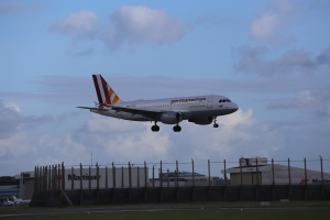 Germanwings Airbus A320-200 beim Start von Frankfurt Airport mit Gras darunter, einem Zaun im Vordergrund, Gebäuden und Bäumen im Hintergrund und einer bewölkten Himmel über.