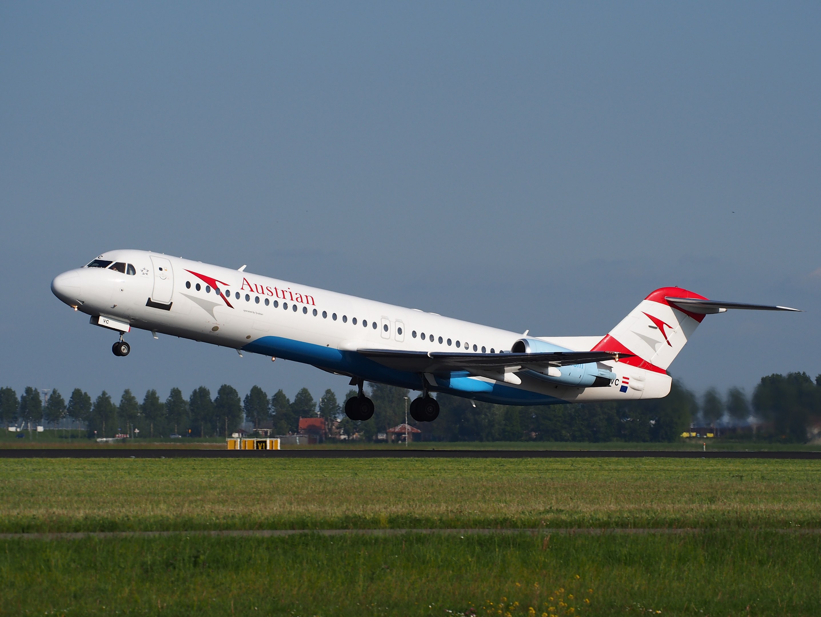 Austrian Airlines Embraer ERJ-190 Flugzeug beim Abheben vom Frankfurt Airport, mit grünem Gras darunter und Bäumen und Gebäuden im Hintergrund unter einem sichtbaren Himmel.