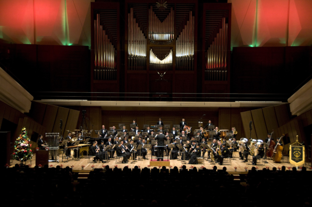 Großes Orchester auf der Bühne mit einem Weihnachtsbaum im Hintergrund, Musiker auf Stühlen sitzend, Dirigent am Pult und Texttafeln auf der rechten Seite.