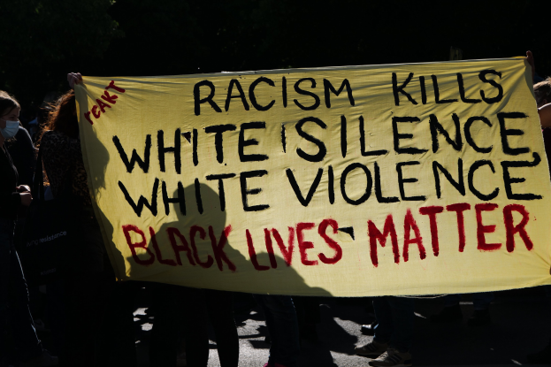 Eine Gruppe maskierter Menschen mit einem Transparent mit der Aufschrift "Rassismus t├Âtet White Silence, White Violence, Black Lives Matter" auf einer dunklen Stra├če.