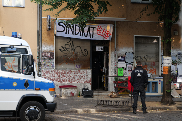 Polizist vor einem Gebäude mit Graffiti, daneben ein Van und ein Baum, Hintergrund zeigt Gebäude mit Fenstern, eine Fahne und andere Gegenstände.