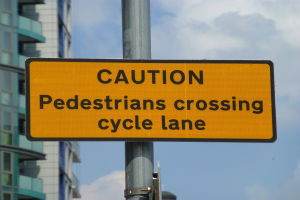Gelbes Warnschild an einem Pfahl vor einem Gebäude, mit einem klaren blauen Himmel im Hintergrund, das 'Fußgänger überqueren Fahrradspur' anzeigt.