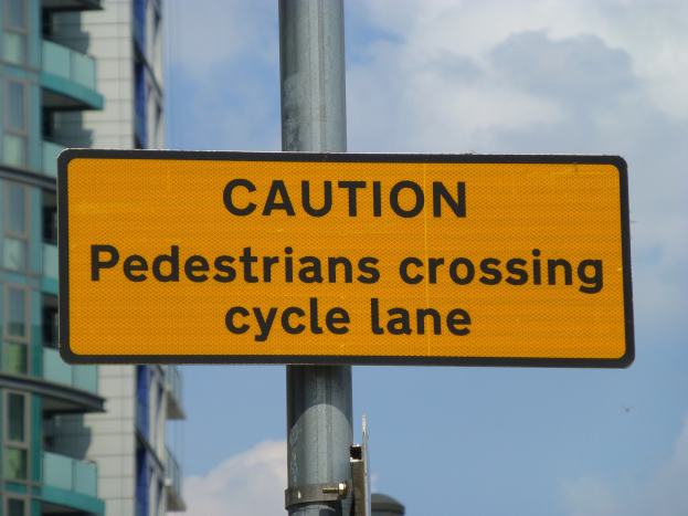 Gelbes Warnschild an einem Pfahl vor einem Gebäude, mit einem klaren blauen Himmel im Hintergrund, das 'Fußgänger überqueren Fahrradspur' anzeigt.