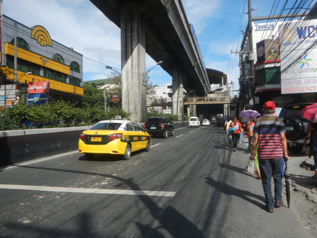 Ein gelbes Taxi fährt auf einer Stadtstraße mit hohen Gebäuden, Passanten auf dem Gehweg, einige halten Schirme, eine Brücke im Hintergrund, bewölkter Himmel und Oberleitungen.