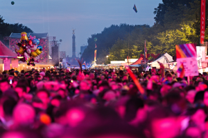 Eine große Menschenmenge auf einem nächtlichen Musikfestival, beleuchtet von bunten Lichtern, umgeben von Zelten, Fahnen, Pfählen und anderen Gegenständen, und im Hintergrund ein Turm, Bäume und einen klaren Nachthimmel.