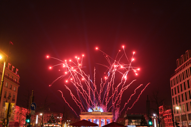 Eine Stadtstraße in Berlin an Silvester, voller Gebäude, Bäume, Laternenmasten, Verkehrszeichen, Zelte und Menschen, mit einem Feuerwerk erleuchteten Himmel im Hintergrund.