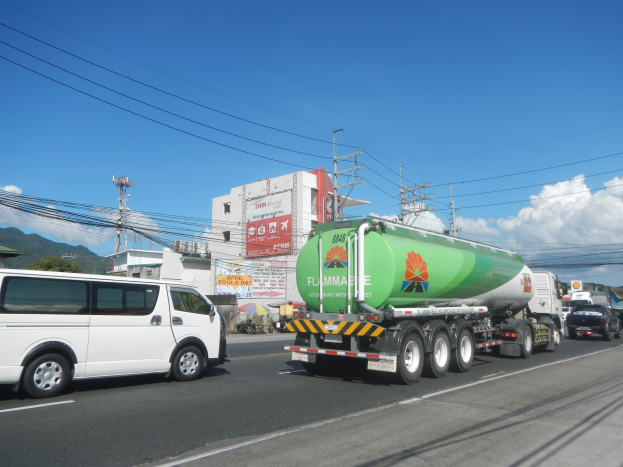 Großer Tanklastzug fährt auf einer Straße neben einem weißen Van mit Strommasten, Gebäuden, Werbetafeln, Bäumen, Hügeln und einem bewölkten Himmel im Hintergrund.
