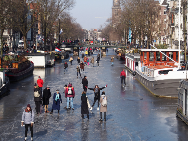 Eine Gruppe von Menschen, die auf einem zugefrorenen Kanal in Amsterdam Eislaufen, umgeben von Booten, Bäumen, Gebäuden mit Fenstern, Laternenmasten, Flaggen und einer Brücke unter einem klaren Himmel.