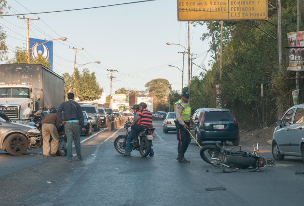 Eine Gruppe von Menschen umringt ein verunglücktes Motorrad am Straßenrand mit mehreren Fahrzeugen, darunter ein Lastwagen, und einer Hintergrundlandschaft mit Bäumen, Pfosten, Laternen und Schildern unter dem Himmel.