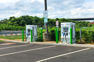 Ladestation für Elektrofahrzeuge auf einem Parkplatz mit umgebender Infrastruktur, Vegetation und einer Brücke im Hintergrund mit wolkenverhangenem Himmel.