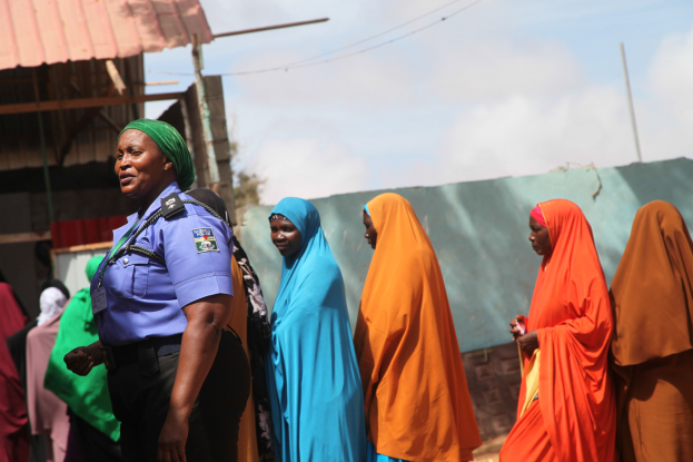 Gruppe von Frauen, die vor einem Gebäude stehen, mit einem Polizeibeamten im Vordergrund, Schuppen, Pfosten, Drähte, Bäume und einen klaren blauen Himmel im Hintergrund.