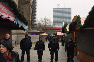 Eine Gruppe von Polizisten in schwarzen Uniformen und Mützen steht vor einem geschmückten Weihnachtsmarkt in Berlin. Im Hintergrund sind Gebäude, Bäume und ein klarer blauer Himmel zu sehen. Auf der linken Seite ist eine Tafel mit Text zu erkennen.