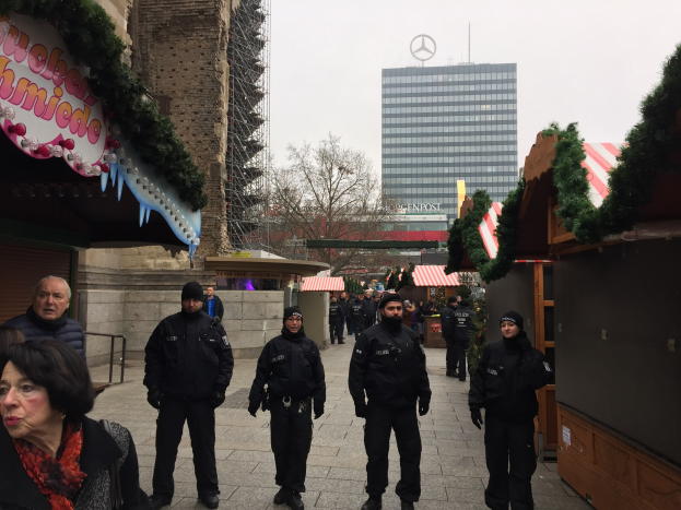 Eine Gruppe von Polizisten in schwarzen Uniformen und Mützen steht vor einem geschmückten Weihnachtsmarkt in Berlin. Im Hintergrund sind Gebäude, Bäume und ein klarer blauer Himmel zu sehen. Auf der linken Seite ist eine Tafel mit Text zu erkennen.