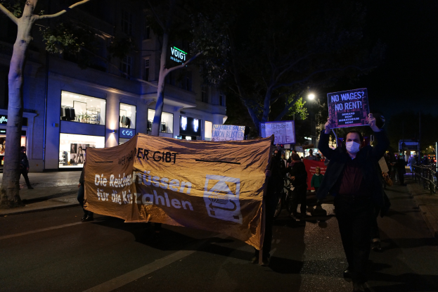 Eine Gruppe von Menschen marschiert nachts auf einer Straße in Berlin, Deutschland, mit Schildern und Plakaten, während Bäume, Gebäude, Laternenpfähle und Fahrräder im Hintergrund zu sehen sind.