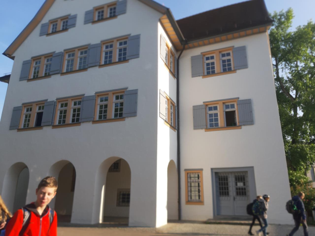 Eine Gruppe von Menschen, einige mit Taschen, geht vor einem großen weißen Gebäude mit Fenstern und Türen, umgeben von Bäumen unter einem klaren blauen Himmel.