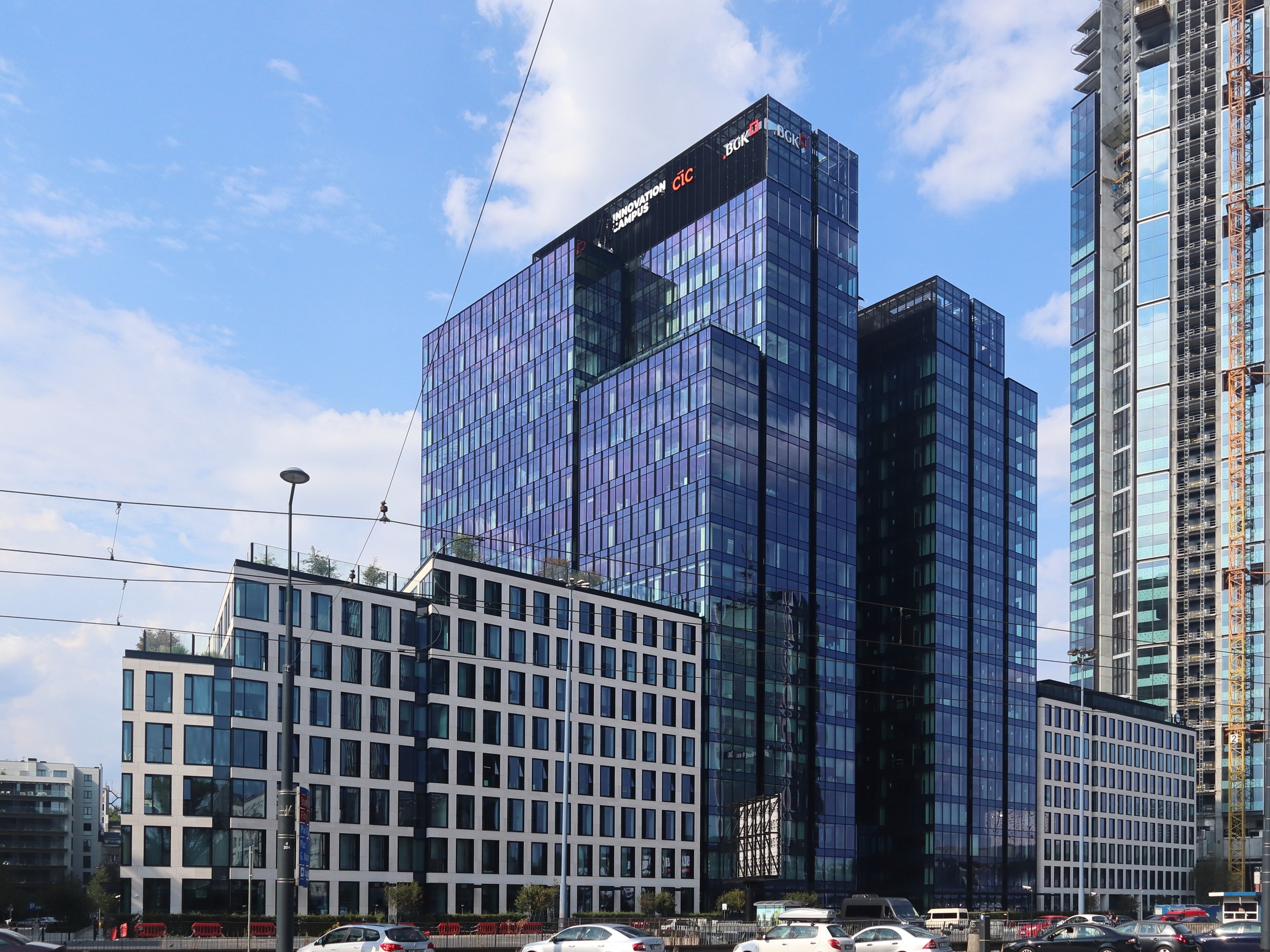 Großes modernes Bürogebäude mit vielen Fenstern, als Deutsche Bank Hauptsitz in Frankfurt, Deutschland identifiziert, umgeben von städtischer Infrastruktur einschließlich Straßenlaternen, Fahrzeugen, Bäumen und einem bewölkten Himmel.