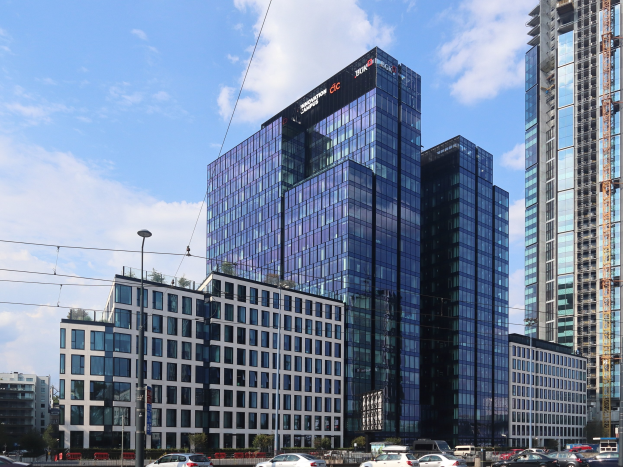 Großes modernes Bürogebäude mit vielen Fenstern, als Deutsche Bank Hauptsitz in Frankfurt, Deutschland identifiziert, umgeben von städtischer Infrastruktur einschließlich Straßenlaternen, Fahrzeugen, Bäumen und einem bewölkten Himmel.