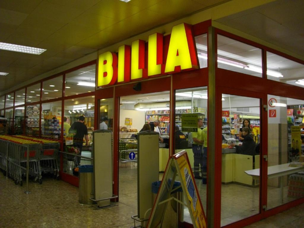 Ein Markt mit einem Eingang in der Mitte, Mülltonnen daneben, eine Reihe von Einkaufswagen auf der linken Seite, Deckenbeleuchtung und einige Menschen, die Produkte anschauen.