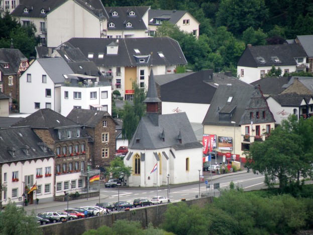 Eine kleine Stadt mit zahlreichen Gebäuden, Bäumen, Fahrzeugen und einer Kirche in der Mitte, mit Pflanzen und Wasser am Bildrand.