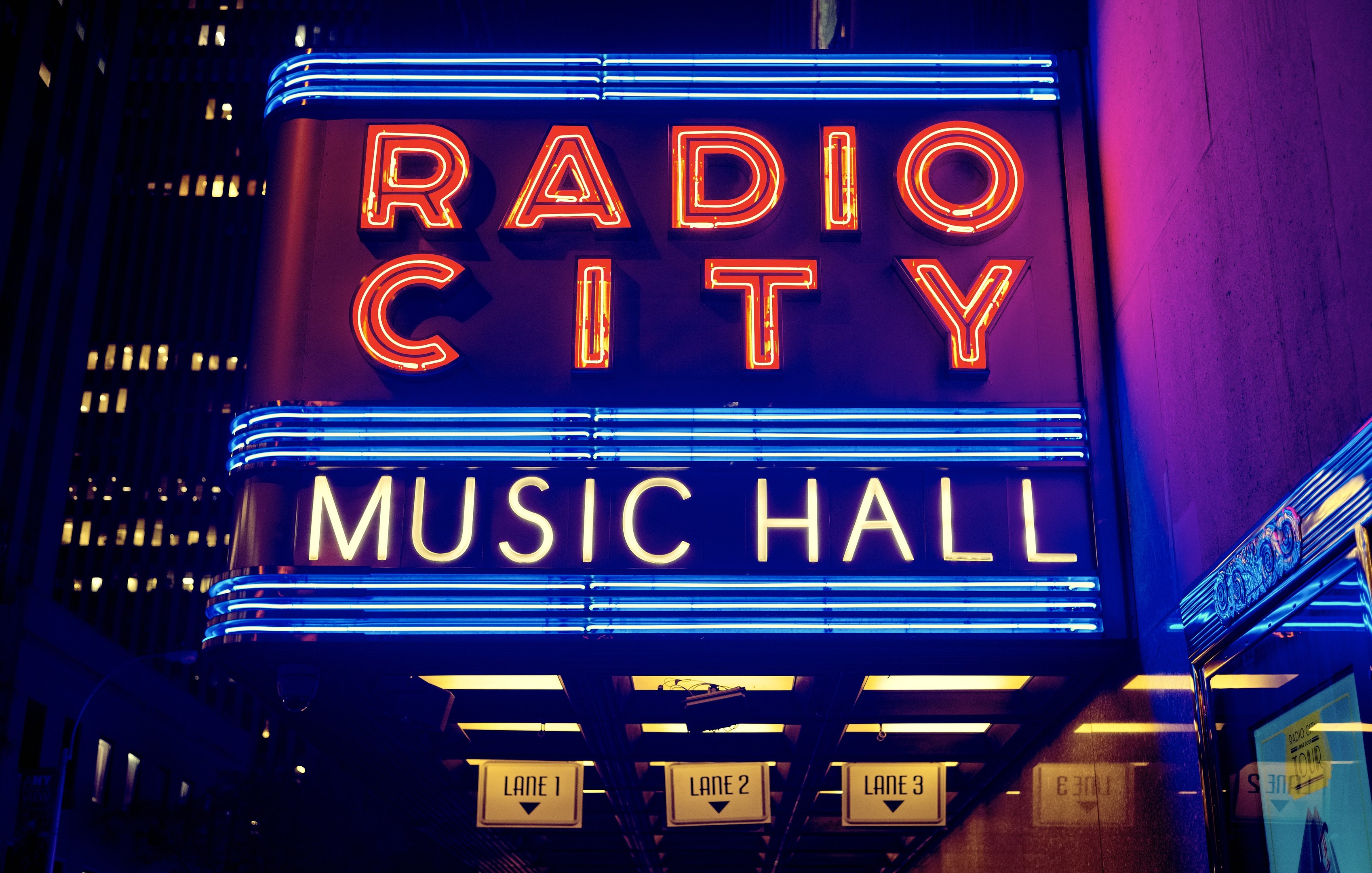 Das erleuchtete Radio City Music Hall in New York City, umgeben von hohen Gebäuden, mit einer 'Radio City'-Leuchtschrift und zusätzlichen Schildern auf der rechten Seite.