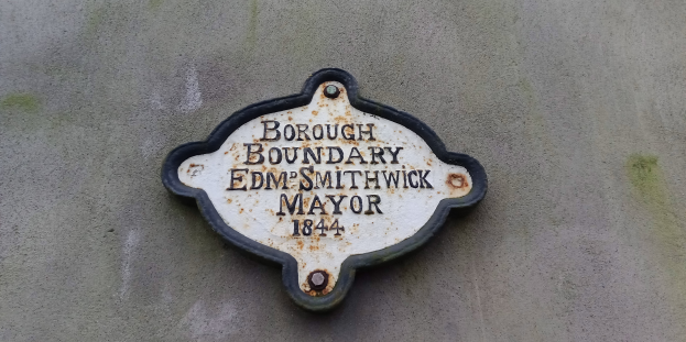 Eine Metalltafel an einem Gebäude mit schwarzem Hintergrund und weißer, fetter, zentrierter Schrift, die "Gemeindegrenze Edm Smithwick Bürgermeister" lautet.