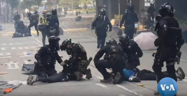 Eine Gruppe von Polizisten in Einsatzausrüstung umringt einen Mann, der auf dem Boden liegt, mit Bäumen, Pfählen, Gebäuden und anderen Gegenständen im Hintergrund.