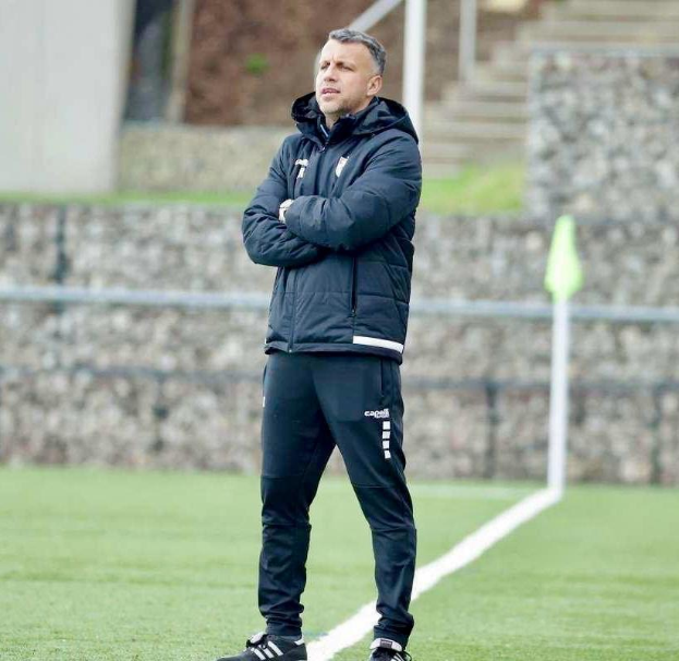 Ein Fussballtrainer auf einem Feld mit verschränkten Armen, der eine schwarze Jacke und Hose trägt, vor einem Zaun, Treppe und Wand während eines Trainings.