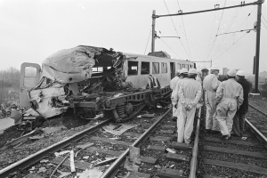 Eine Schwarz-Weiß-Szene eines Zugunglücks mit zwei Zügen auf den Schienen, umgeben von Menschen in Helmen, mit Strommasten, Bäumen, Gebäuden und einem klaren Himmel im Hintergrund.