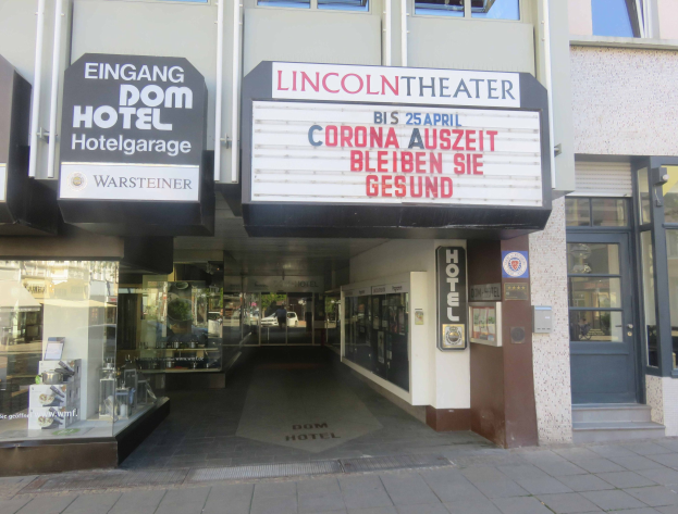 Außenansicht des Lincoln Theaters in Berlin, Deutschland, mit Glasfenstern und -türen sowie einer Tafel mit Text und einem Inneren, das eine pulsierende Stadtlandschaft suggeriert.