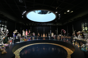 Ein großer Raum voller verschiedener Spielzeuge, darunter Skelette, die auf dem Boden angeordnet sind, mit Schädeln an den Wänden und Lichtern an der Decke, die eine festliche Stimmung für den Tag der Toten schaffen.