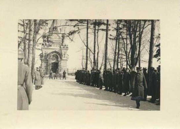 Schwarz-weißes Foto einer Gruppe von Menschen, die vor einem Gebäude mit einem Torbogen im Hintergrund und Bäumen an der Straße stehen, mit dem Text "Die Trauerfeier für die Opfer der Berliner Mauer" unten.