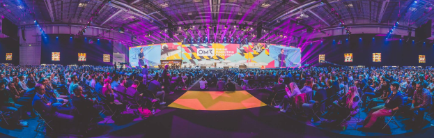 Eine große Menschenmenge, die auf Stühlen sitzt und einer hell erleuchteten Bühne gegenübersteht, auf der mehrere stehende Personen zu sehen sind, wahrscheinlich bei einem Online-Marketing-Event.