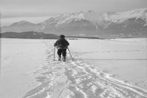 Eine Person Skilanglauf im Schnee mit Bergen im Hintergrund, die eine Jacke, eine Mütze und Handschuhe trägt und Ski-Stöcke hält.