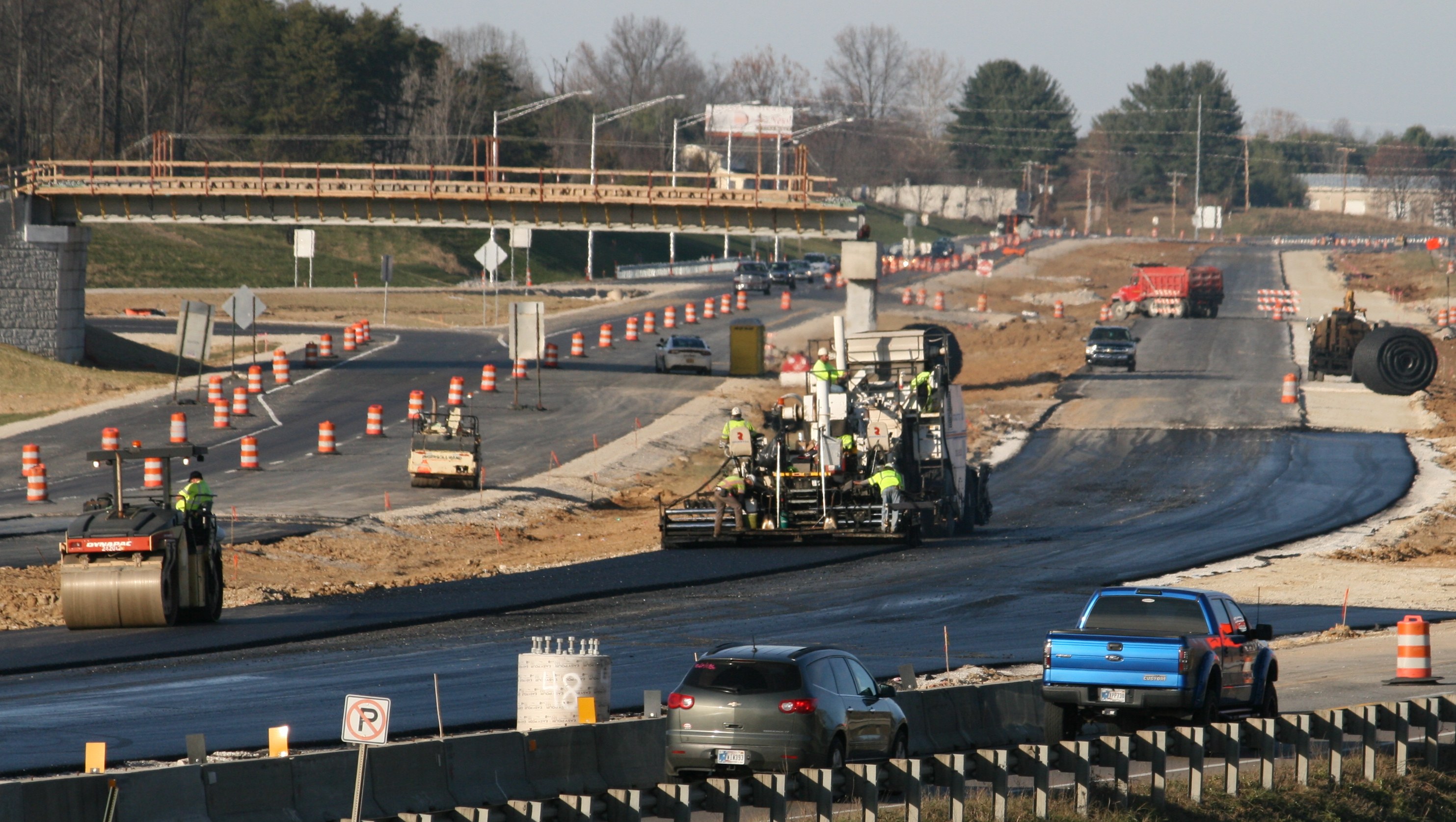 Baustelle mit Fahrzeugen auf der Straße, Verkehrskegel, Hinweisschilder, Pfosten, eine Brücke, Bäume und einen klaren blauen Himmel.